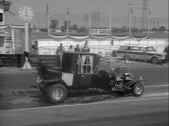 Vintage dragster on a racetrack near the pit area, with cars and spectators in the background.