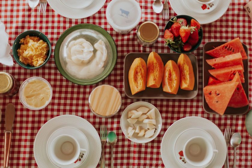 Café da manhã servido sobre uma toalha de mesa xadrez vermelha, com panquecas, waffles, frutas, iogurte e xícaras de café.