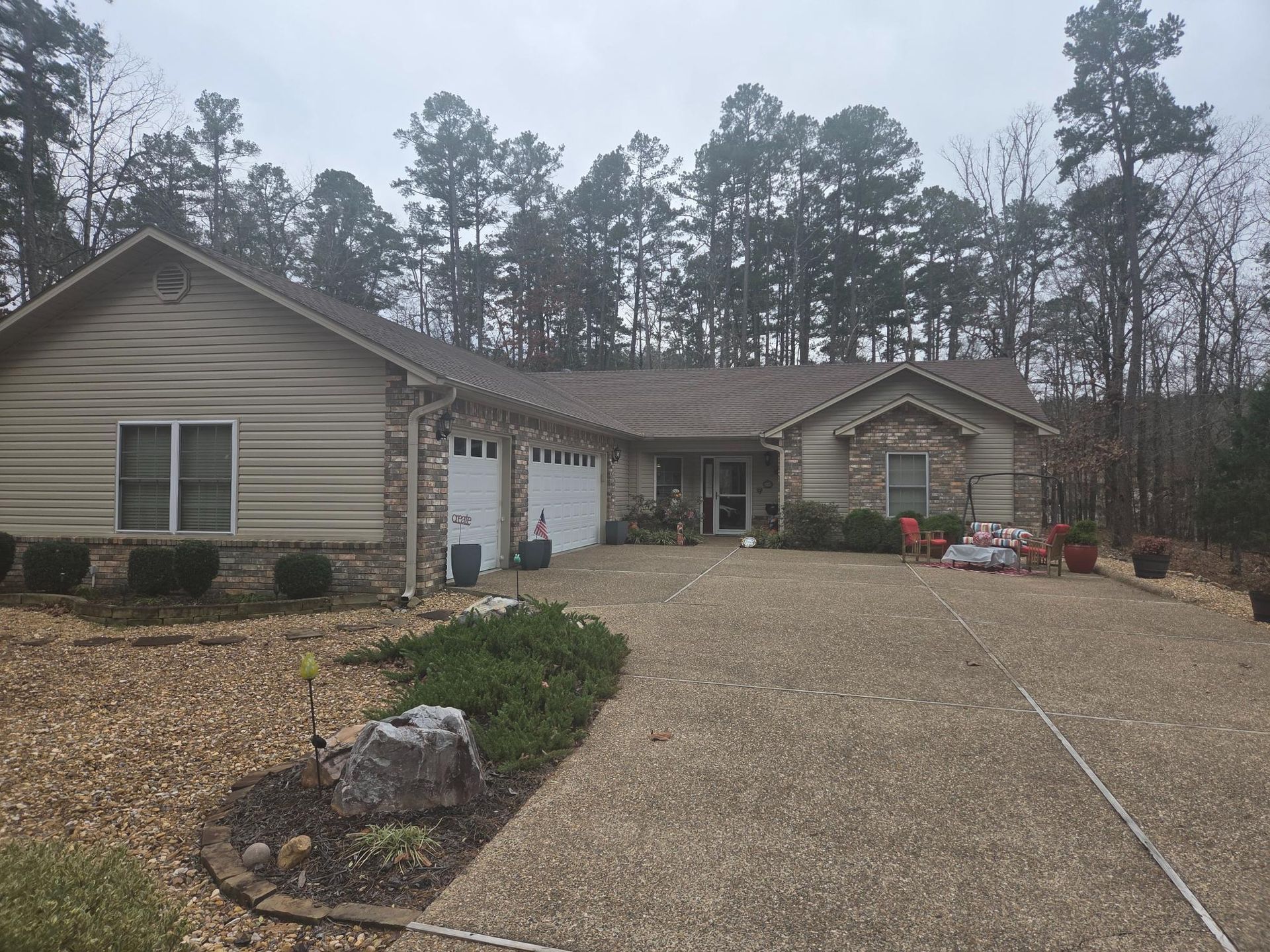 Tan and stone-faced ranch house with a gravel driveway, surrounded by trees and a cloudy sky.