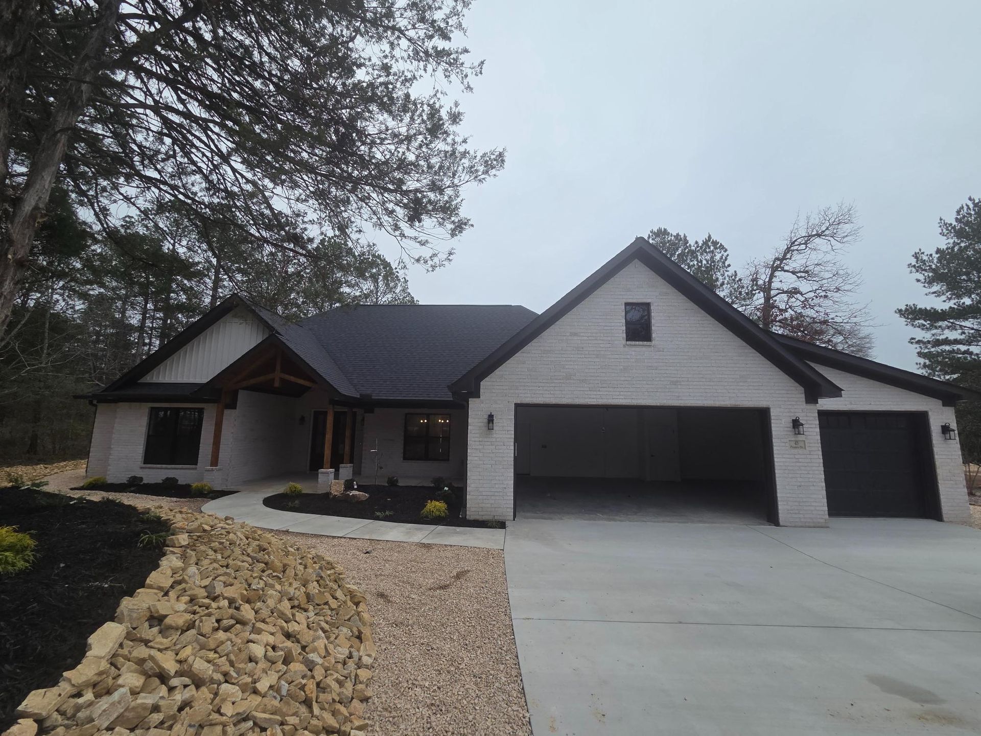A light brick-faced house with a black roof and a two-car garage, gray driveway, and gravel landscaping.