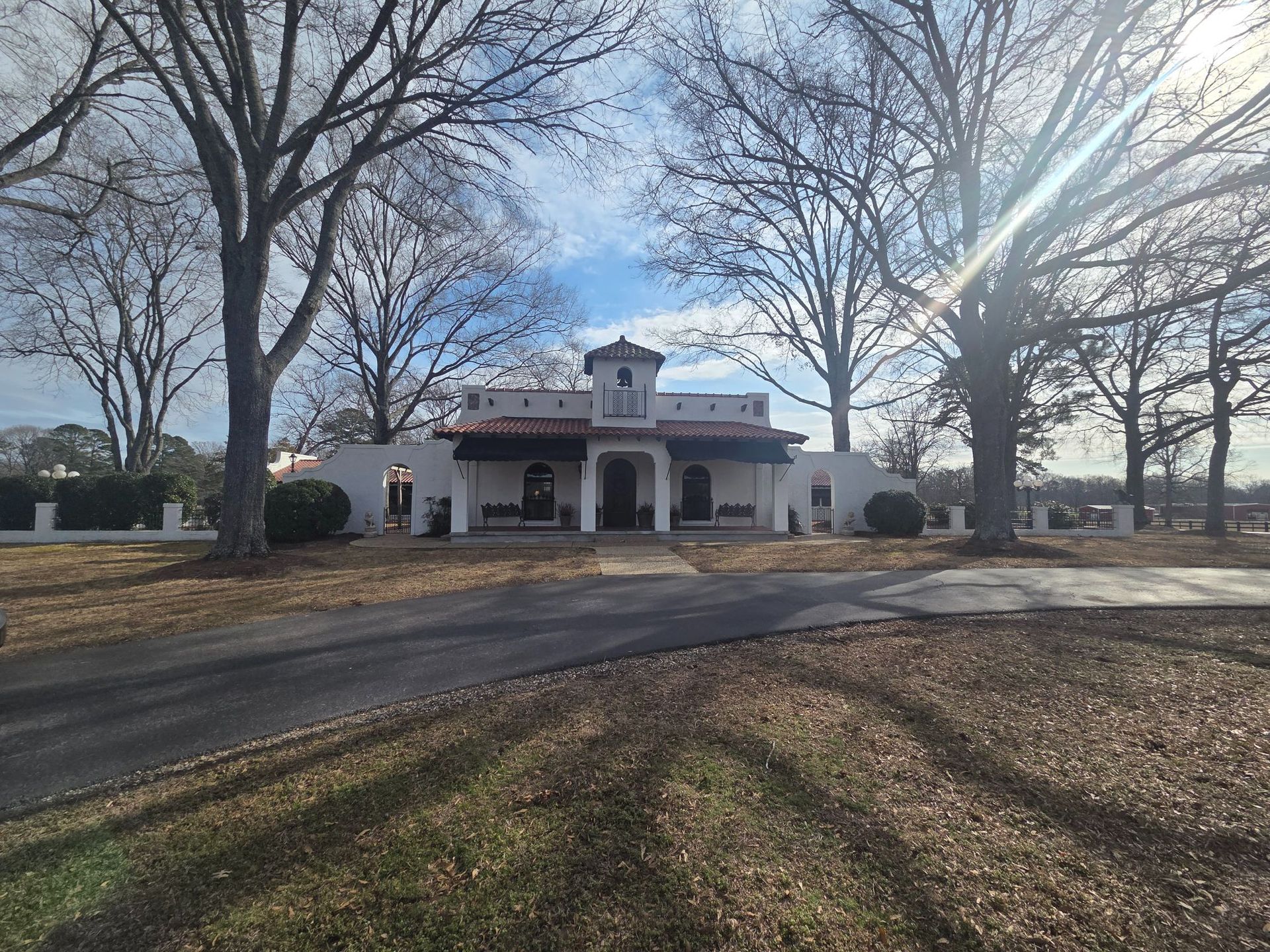 White stucco building with red tile roof, arched windows, and a driveway lined with trees.