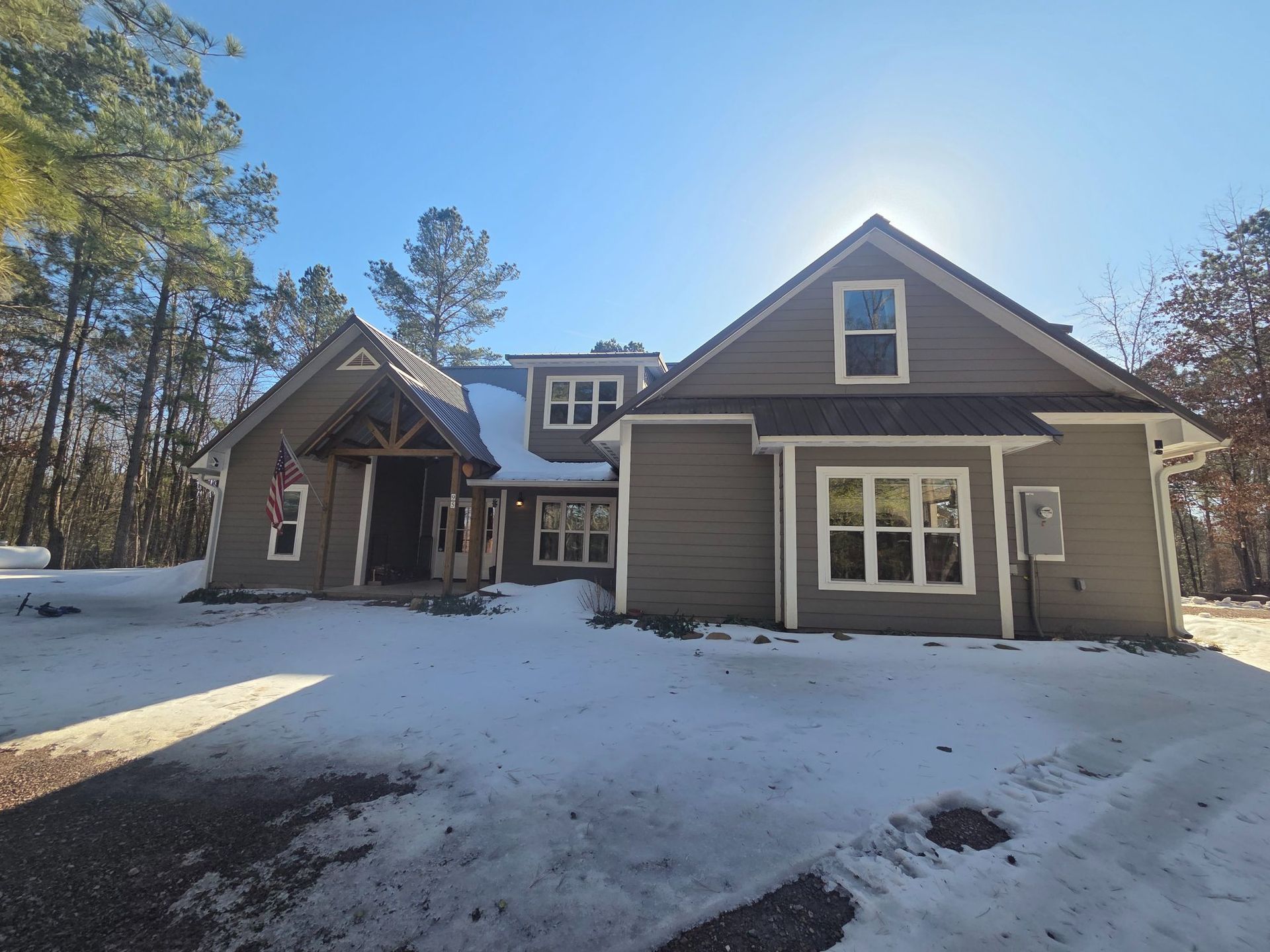 House with snow-covered lawn. Gray siding, white-trimmed windows, porch with flag. Sunny winter day.
