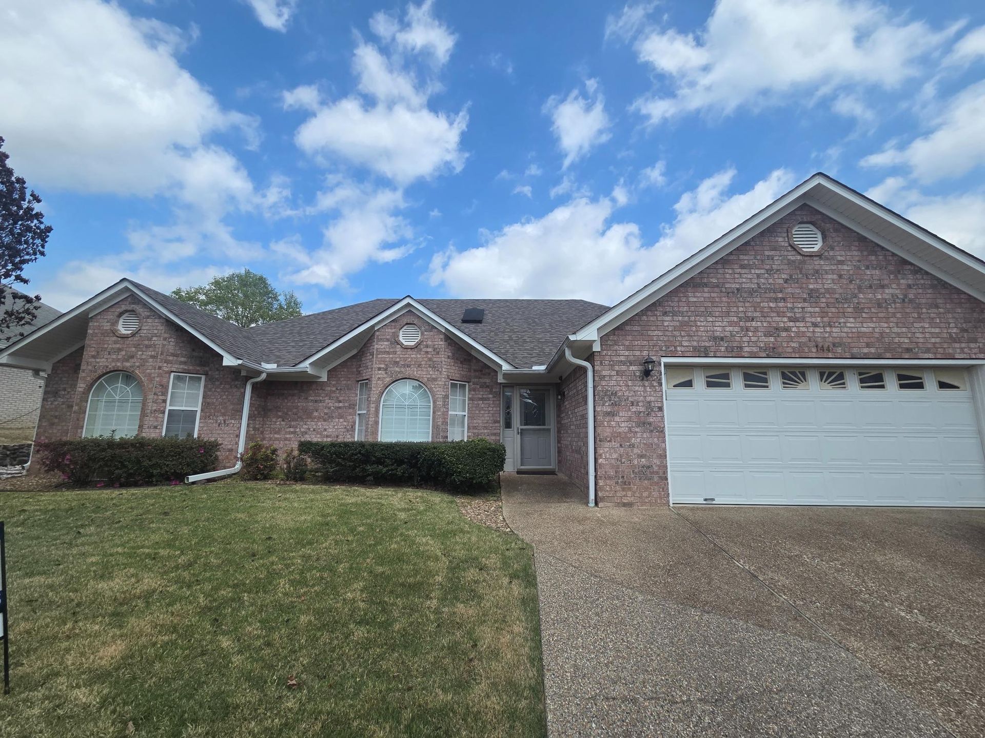Single-story brick house with a front-facing garage, gravel driveway, and lawn under a blue sky with scattered clouds.