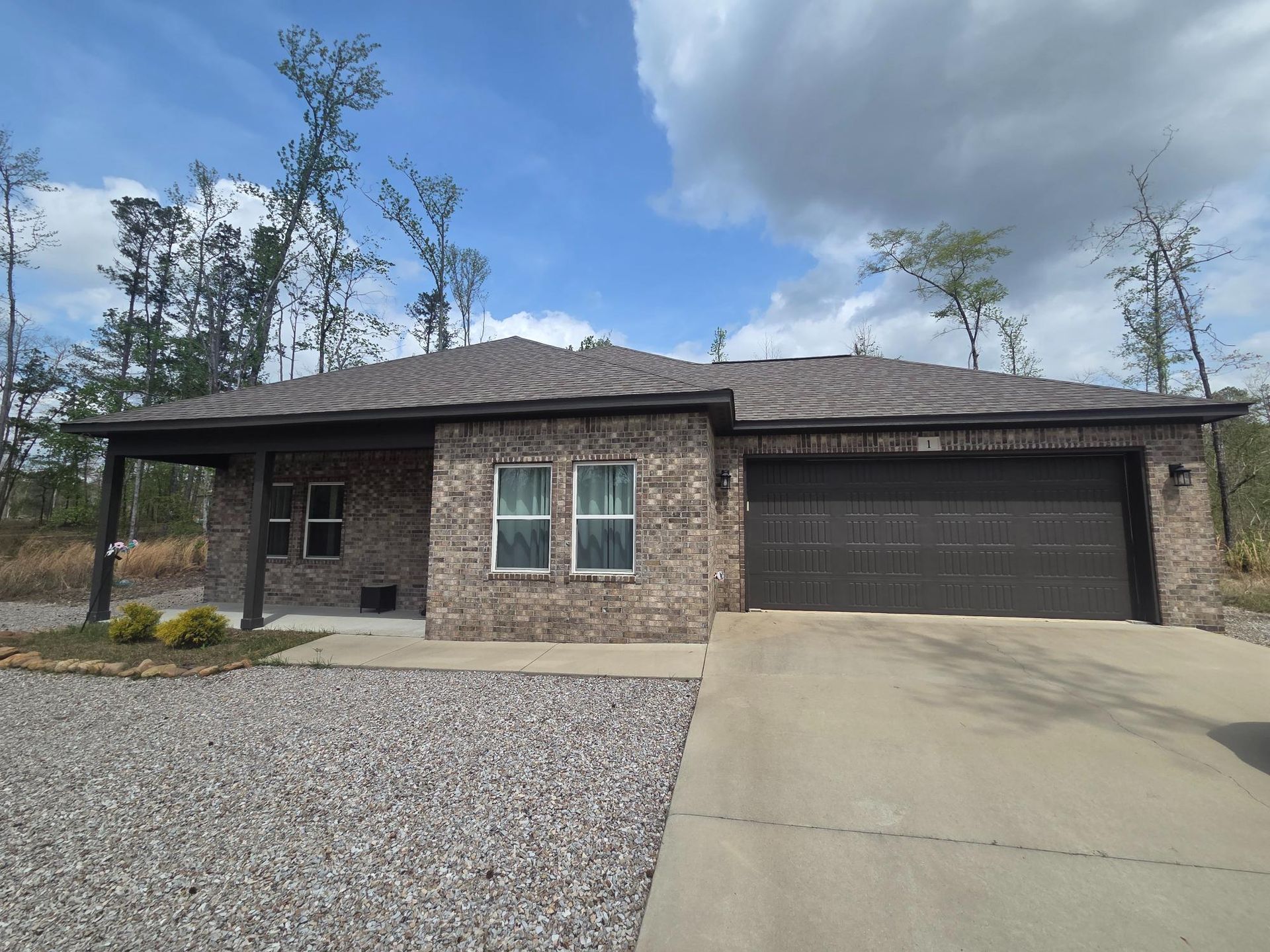 A single-story, tan brick house with a dark roof and garage, fronted by a concrete driveway and gravel yard.