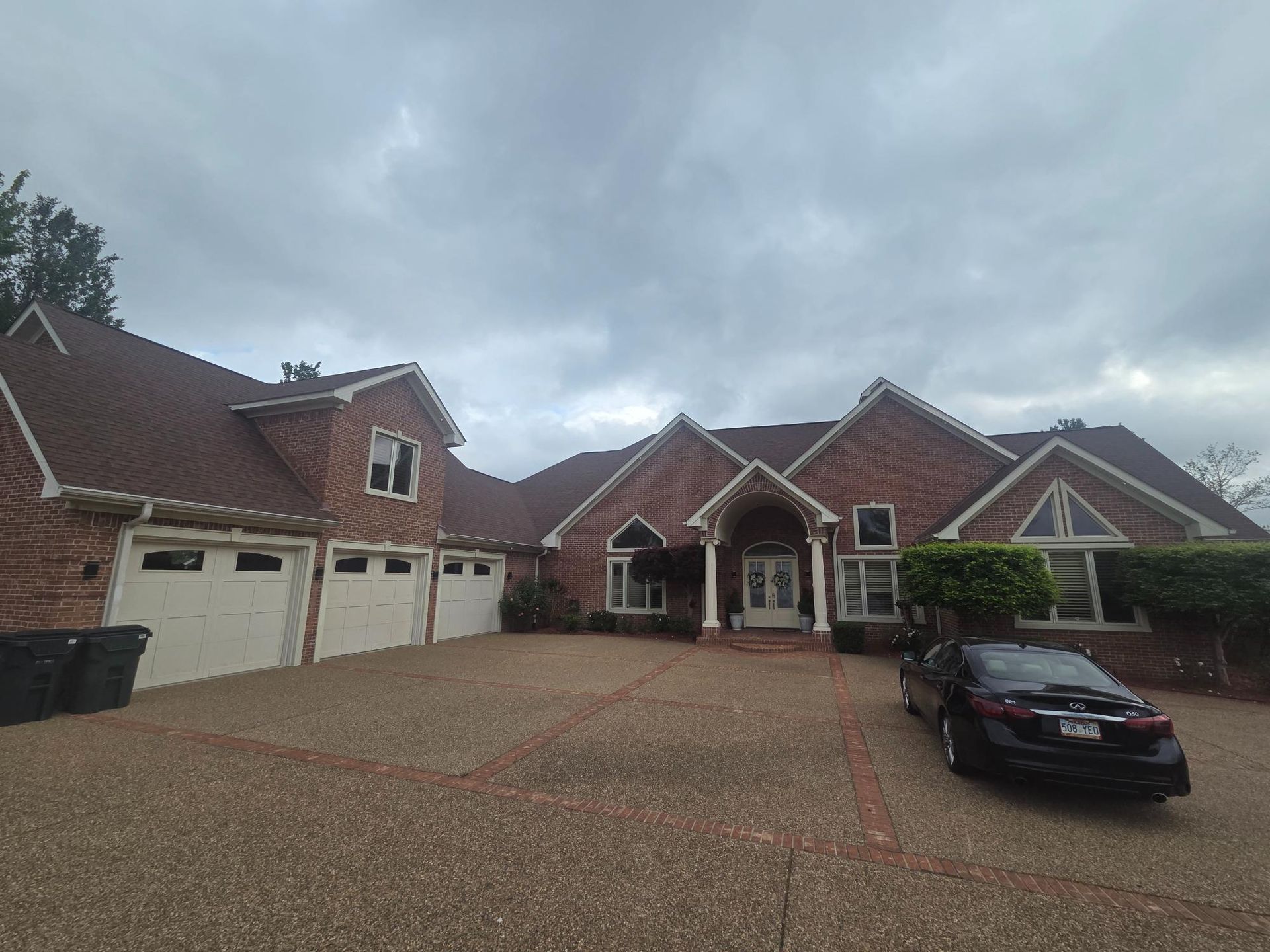 A large brick home with a three-car garage and a wide stone driveway under a cloudy sky, with a car parked in front.