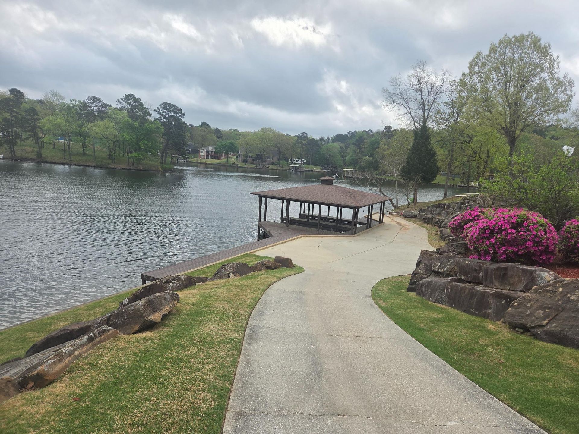 A paved path leads toward a wooden boat dock on a lake, surrounded by green trees, grass, and a bright pink azalea bush.
