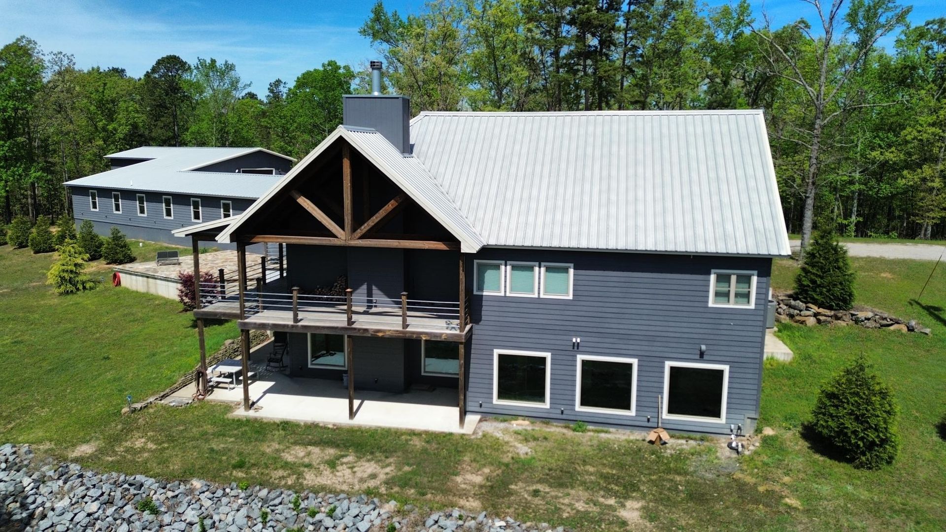 A dark blue, multi-level house with a light metal roof and a spacious wooden deck, set against a backdrop of green trees.