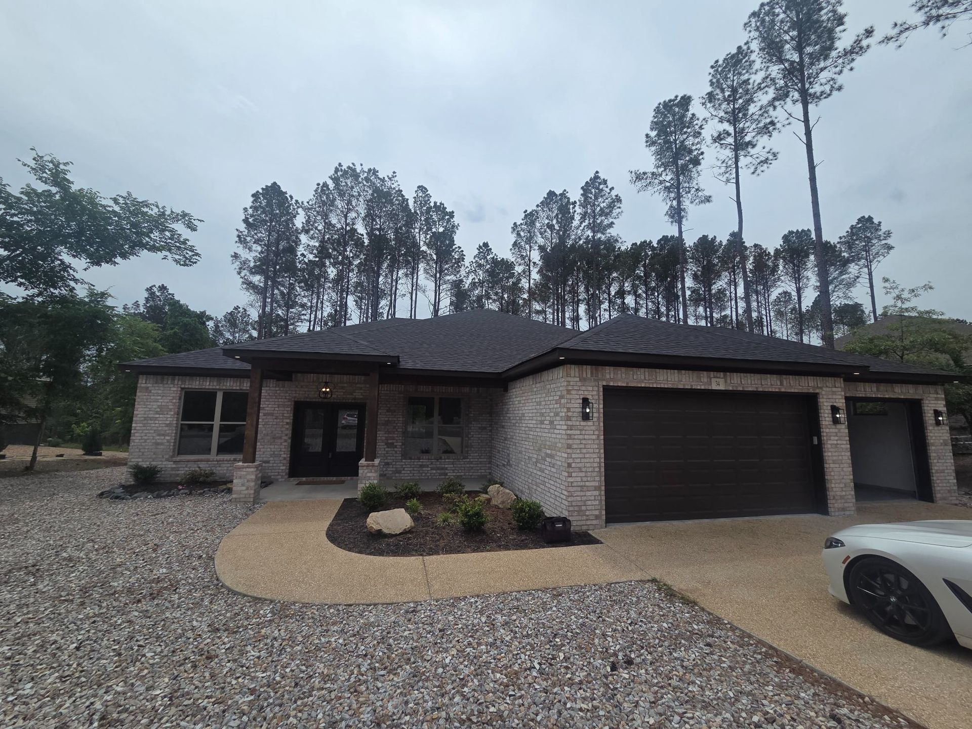 Single-story house with stone facade, dark roof and garage, on a gravel driveway with tall trees behind.