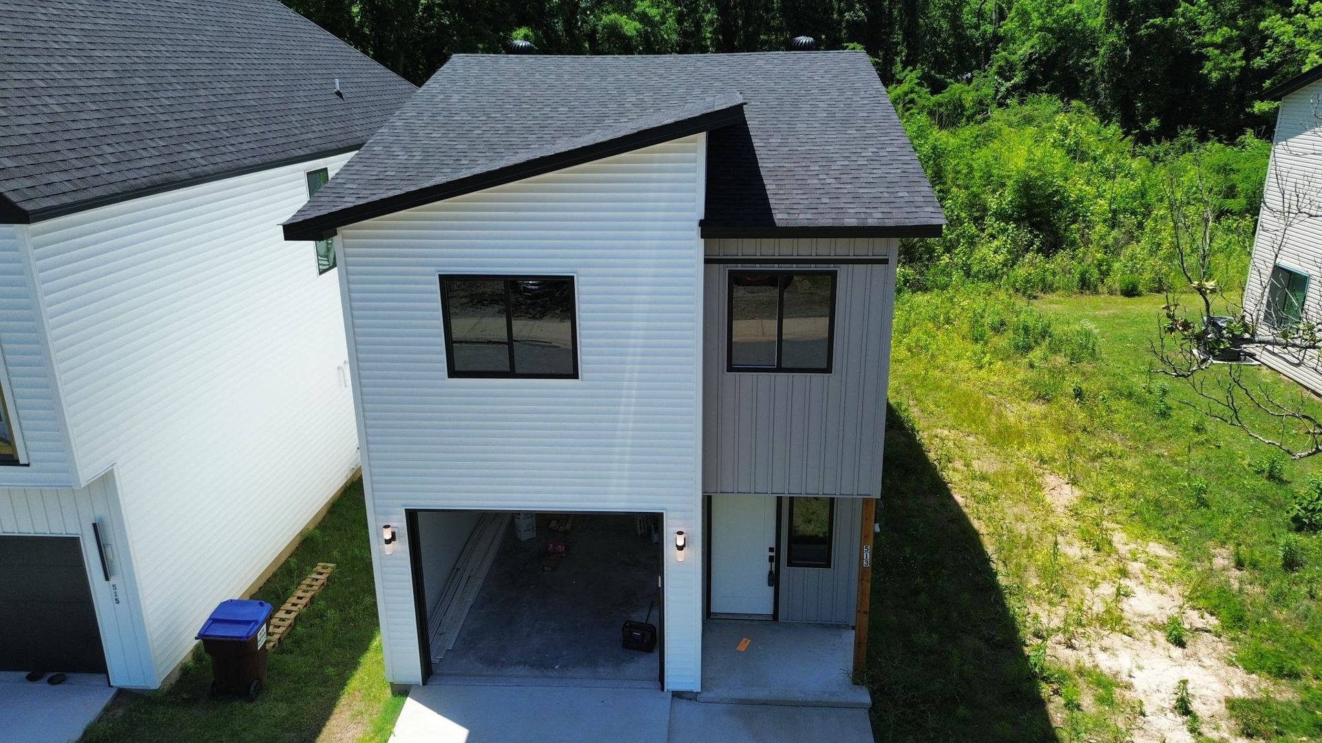 Small white two-story house with a garage, black roof, and grassy yard viewed from above.