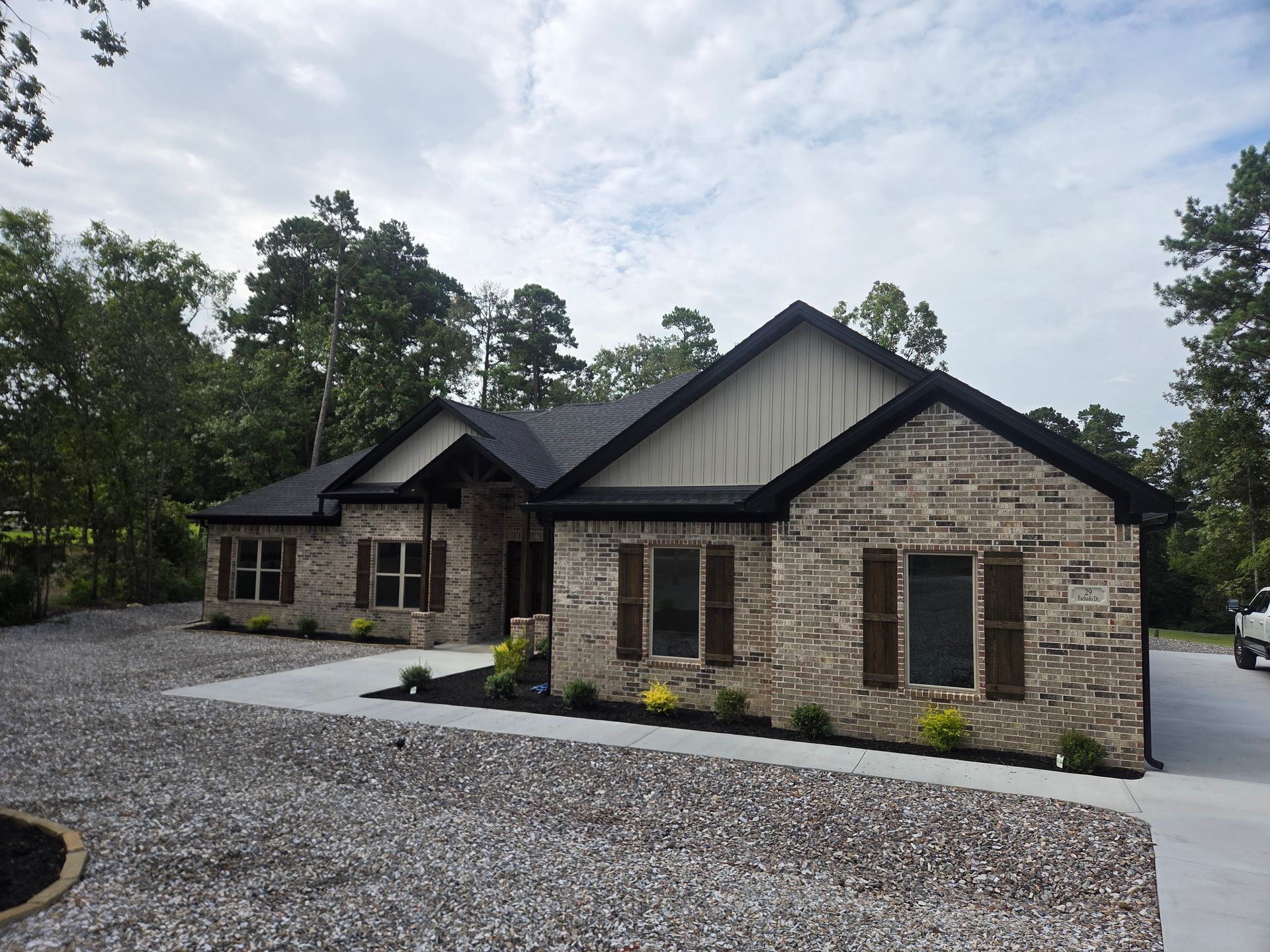 Stone house with dark roof and gravel driveway, surrounded by trees under a cloudy sky