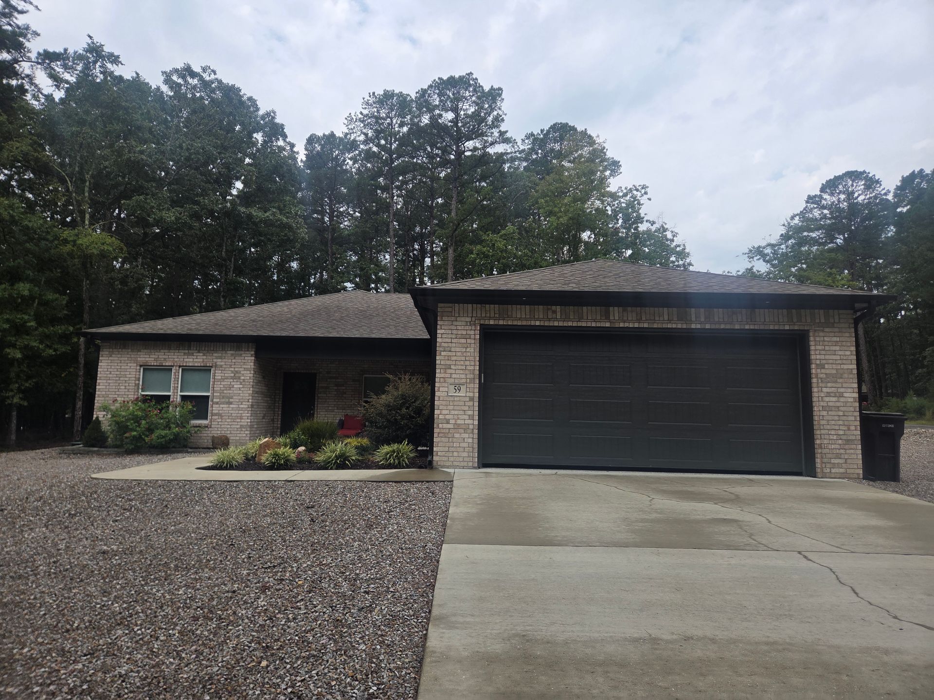 Single-story brick house with a dark garage door and gravel driveway, surrounded by trees