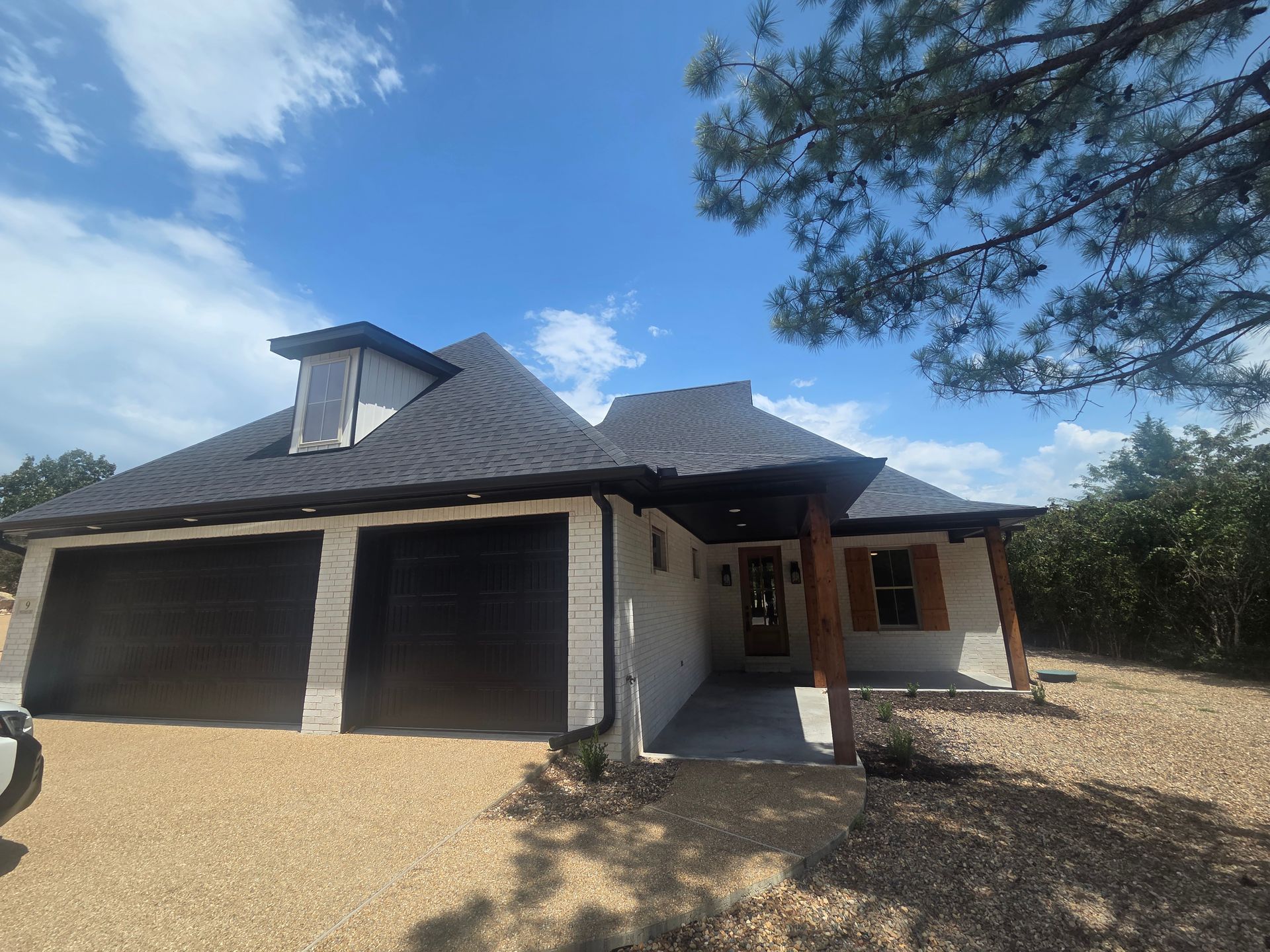 New house with dark roof, double garage, and covered front porch on a gravel lot under a blue sky