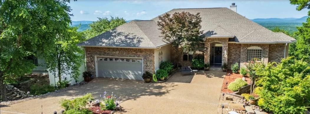 A house with a brick facade and a gray garage door, surrounded by trees and a long driveway.
