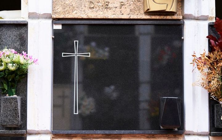 Una bara di legno con composizioni floreali e candele accese in un ambiente sobrio.