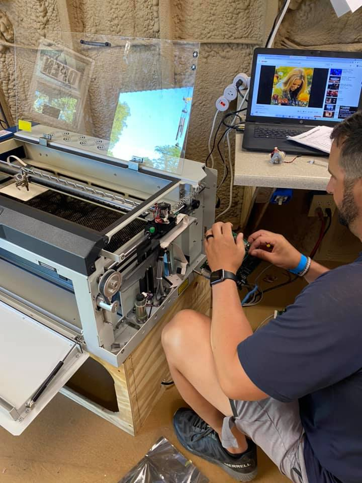 A person sits on the floor, adjusting a desktop laser cutter next to a table with a laptop.