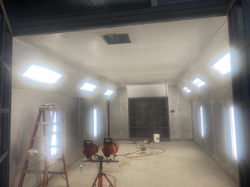 An interior view of a bright, white-walled paint booth with ceiling lights, a ladder, and portable floor lights.