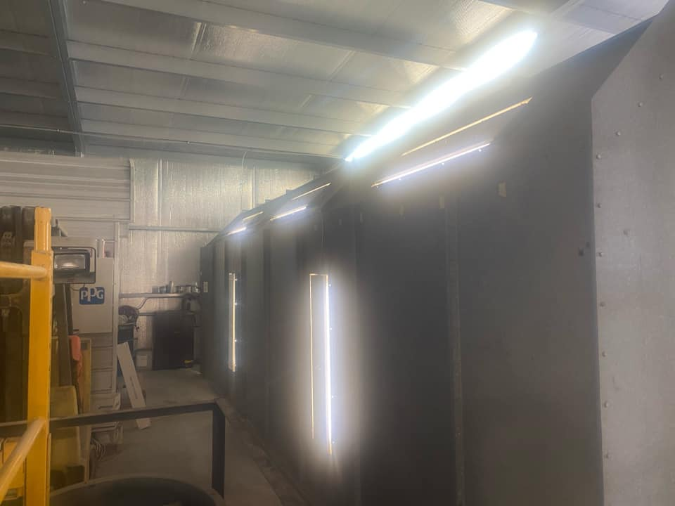 An industrial space with a dark, long spray booth illuminated by bright, vertical and angled LED light strips.