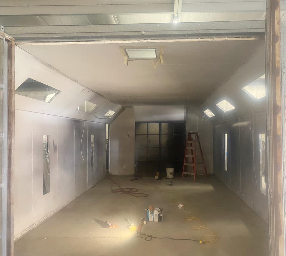 An empty automotive spray paint booth with white walls, recessed ceiling lights, a wooden ladder, and floor equipment.