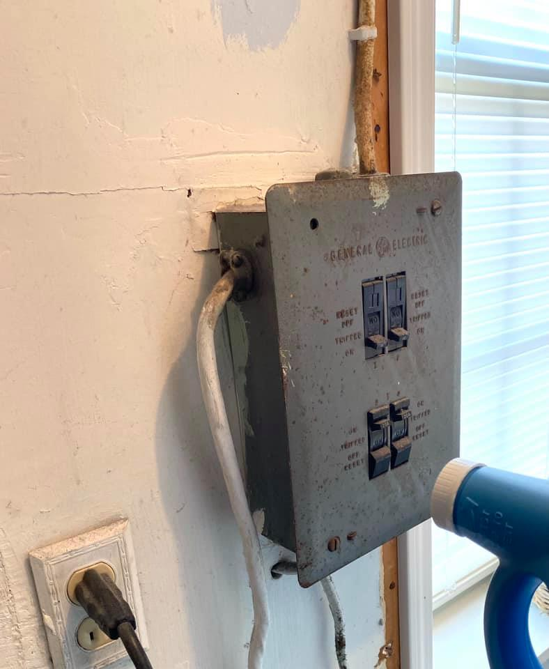 An old gray metal General Electric electrical breaker box mounted on a wall with wires extending from its side and top.