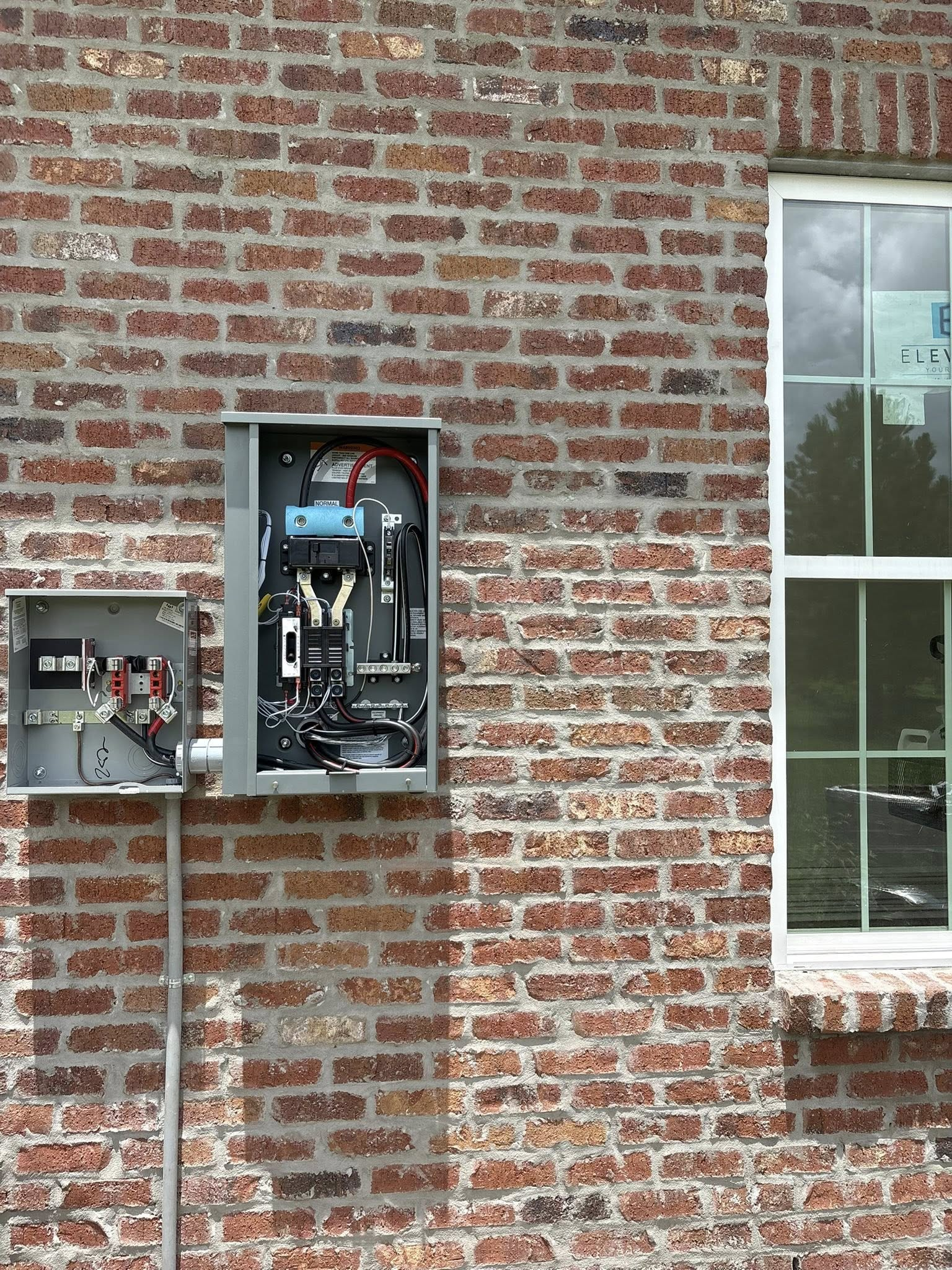 Electrical service equipment consisting of two gray metal boxes mounted on a rustic red brick wall beside a window.