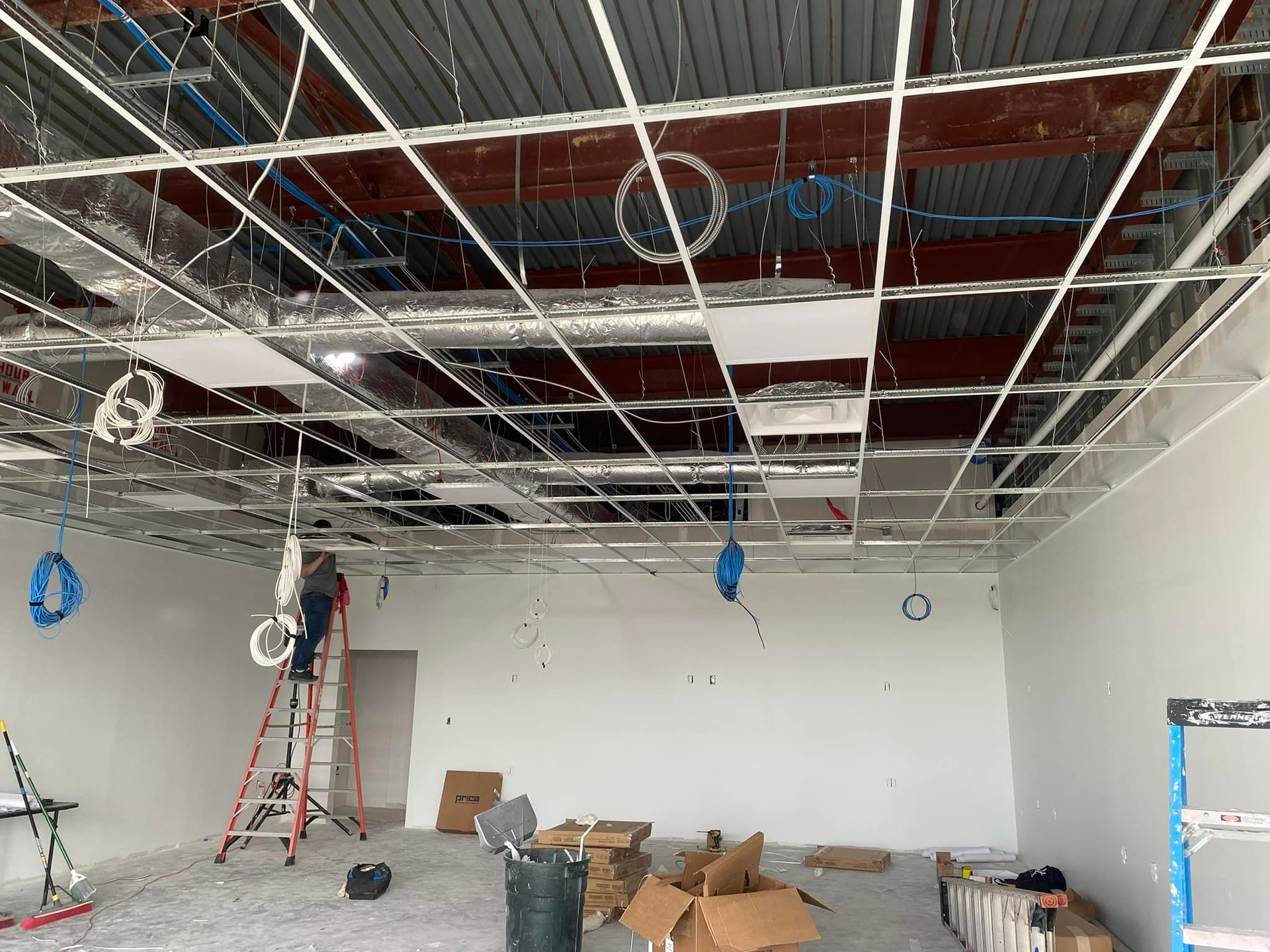 Interior construction site showing a suspended ceiling grid, HVAC ducts, wiring, and a ladder against a white wall.
