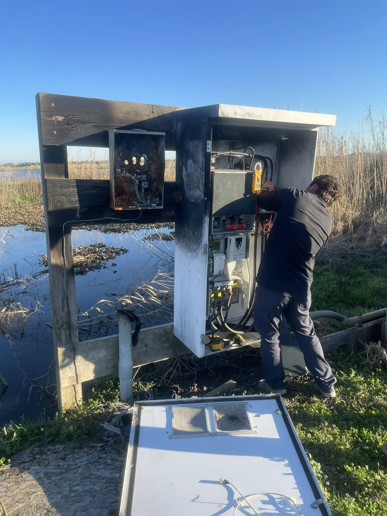 A person in dark clothing inspects a severely fire-damaged outdoor electrical utility box located near a body of water.