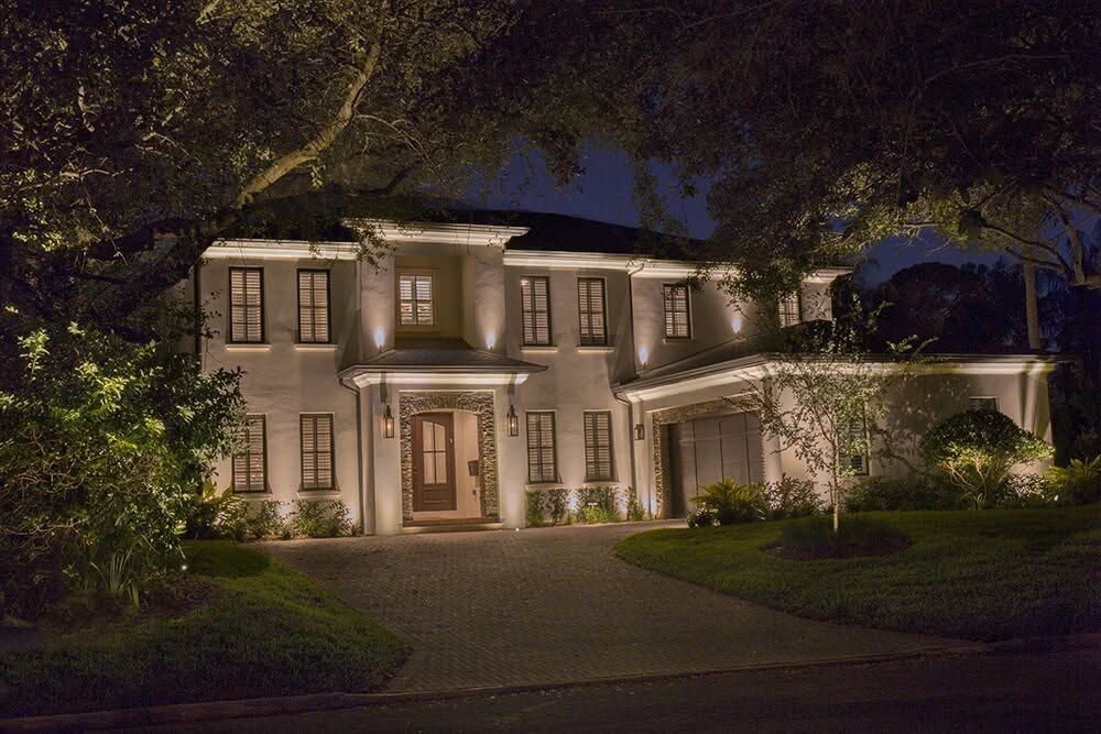 Illuminated two-story house at night with lit windows, front porch, and curved driveway framed by trees