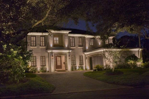Illuminated two-story house at night with lit windows, front porch, and curved driveway framed by trees