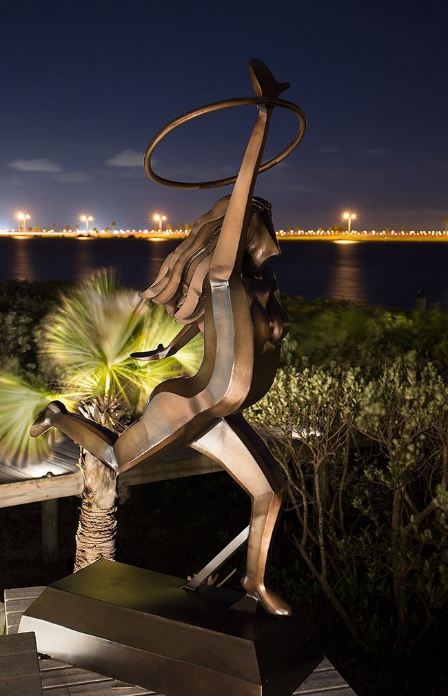 Metal sculpture of a dancing figure lit at night beside water, with city lights in the background