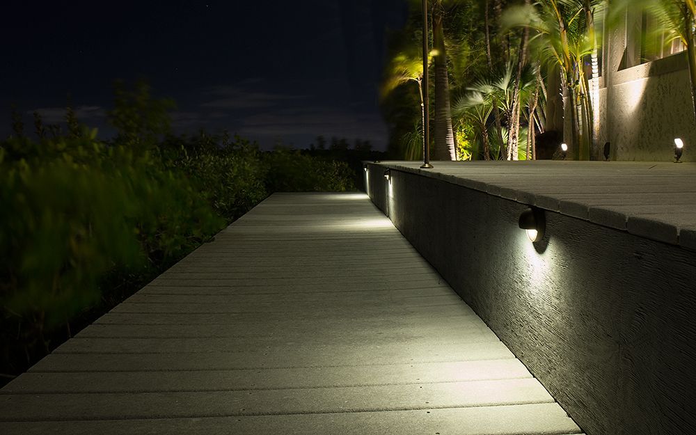 Illuminated wooden walkway beside a raised wall with palm trees at night
