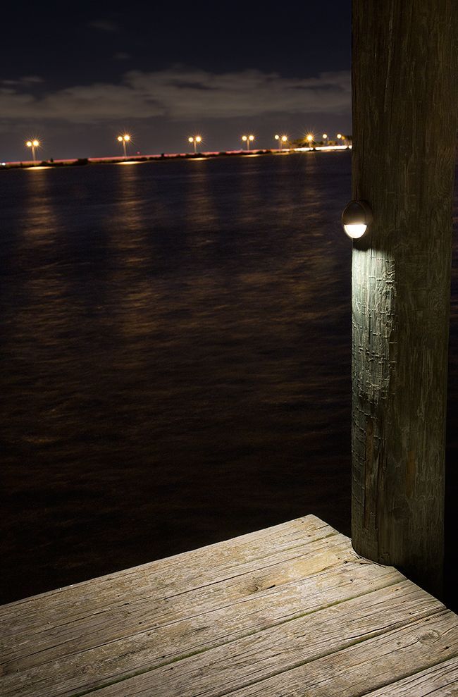 Night waterfront with lit distant lights, dark water, and a wooden dock in the foreground
