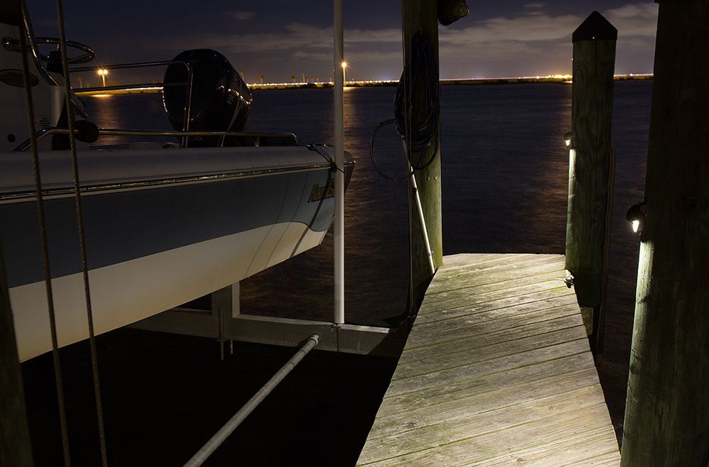 Nighttime dock beside a boat, with lit wooden planks leading to dark water and distant lights.