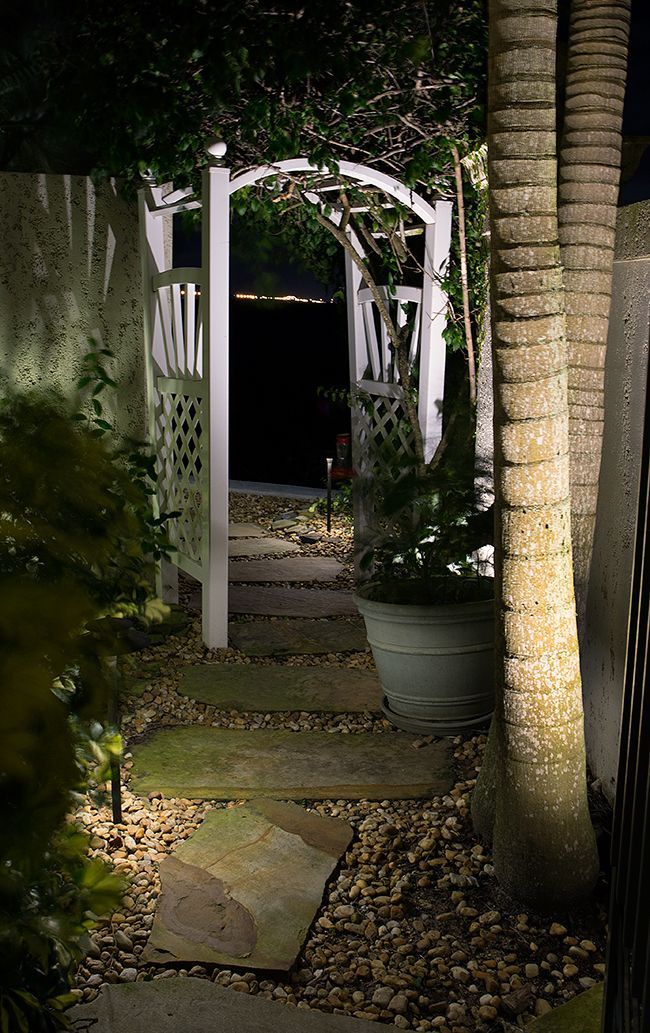 Lit garden path leading to a white archway at night, with palm trees and potted plants.