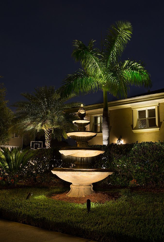 Lit four-tier fountain in a landscaped yard at night, beside palm trees and a house