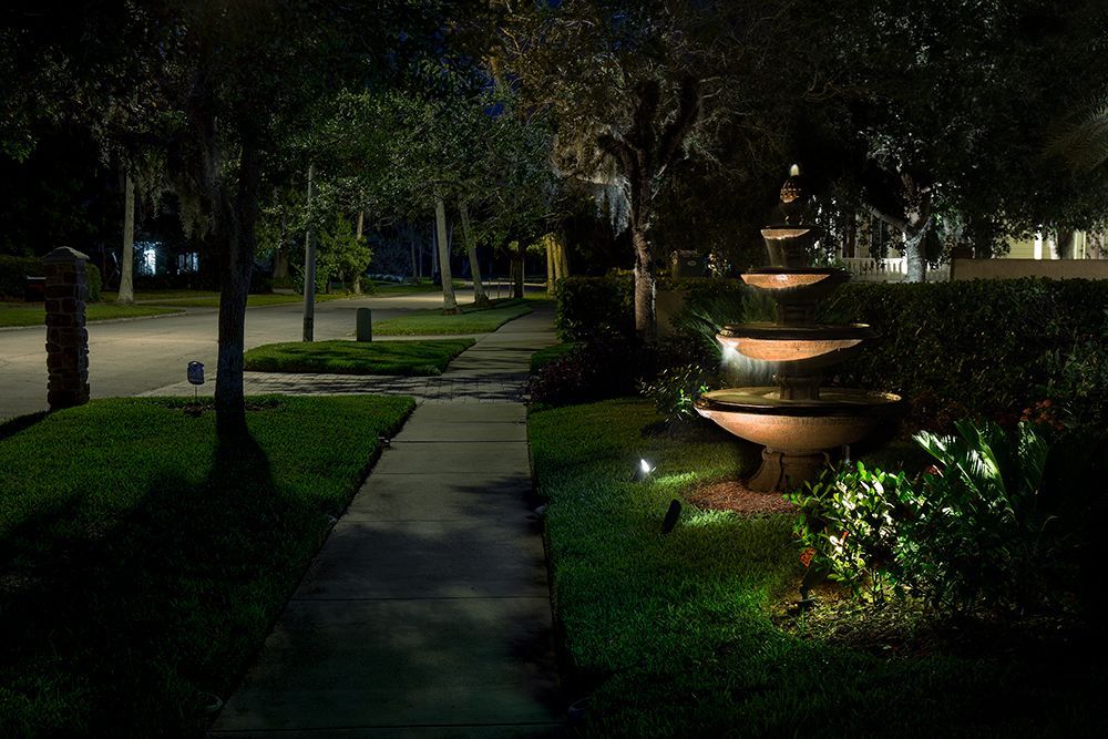Nighttime garden path beside a lit tiered fountain, with trees and hedges in the background
