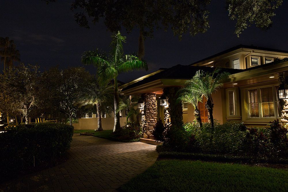 Nighttime view of a warmly lit house with palm trees, a curved walkway, and landscaped garden