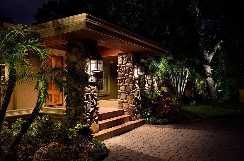 Warmly lit house entrance at night with stone pillars, steps, palm plants, and a paved driveway