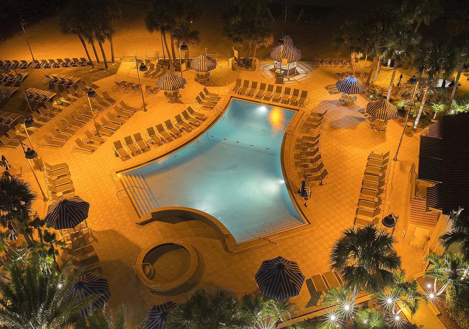 Nighttime resort pool with blue water, orange lighting, and rows of lounge chairs surrounding it
