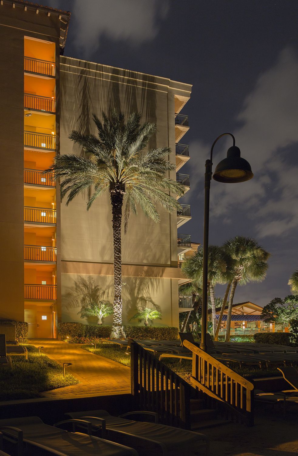 Nighttime hotel courtyard with lit palm tree, building facade, and stairway railing under cloudy sky