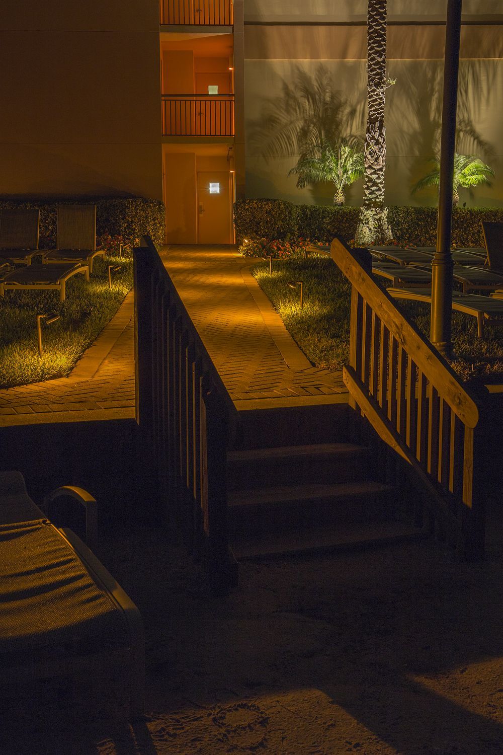Nighttime walkway leading to a lit apartment entrance, with trees and warm orange lighting.