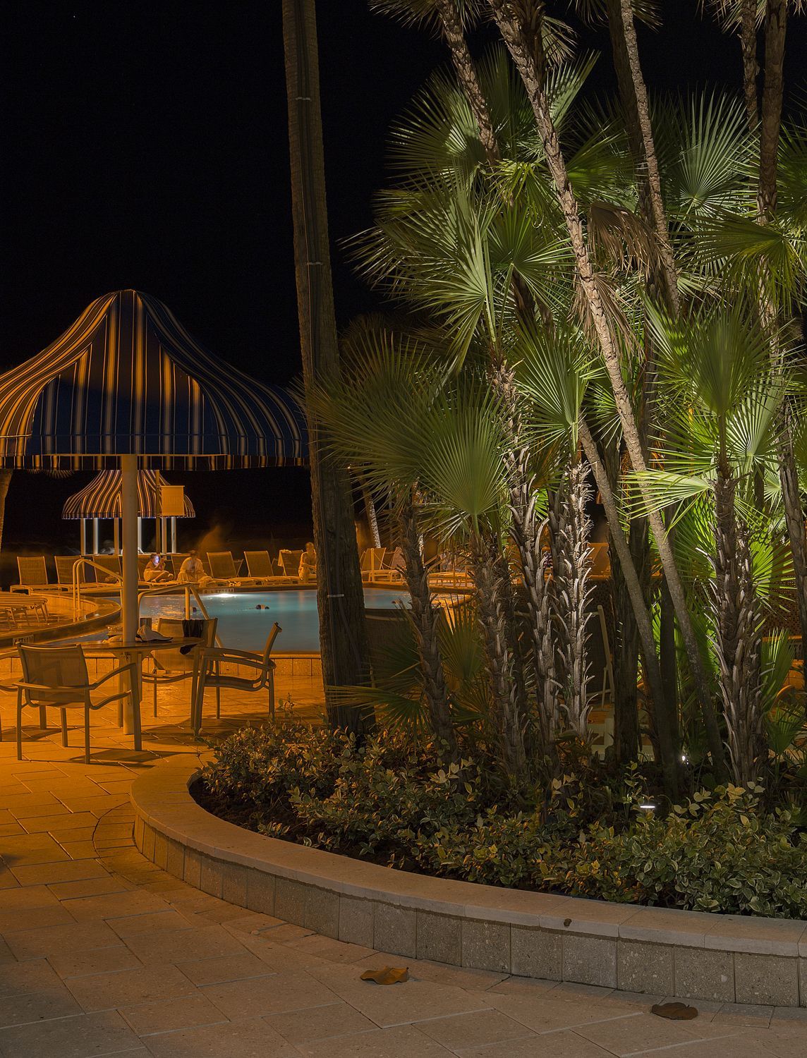Nighttime resort patio with striped cabana, tables, and palm trees lit by warm lights