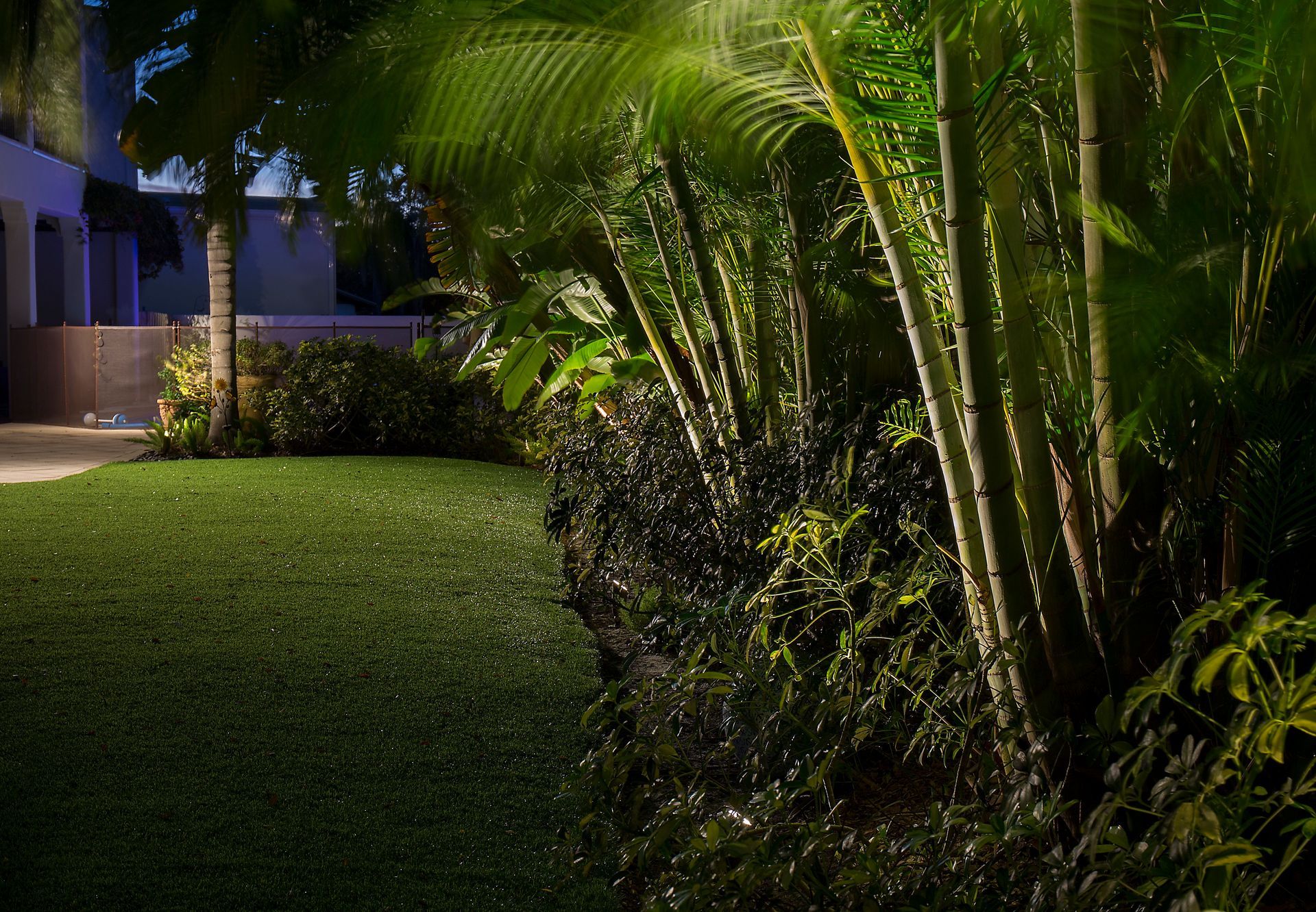 Dimly lit garden path beside a house with lush green lawn and palm trees at night