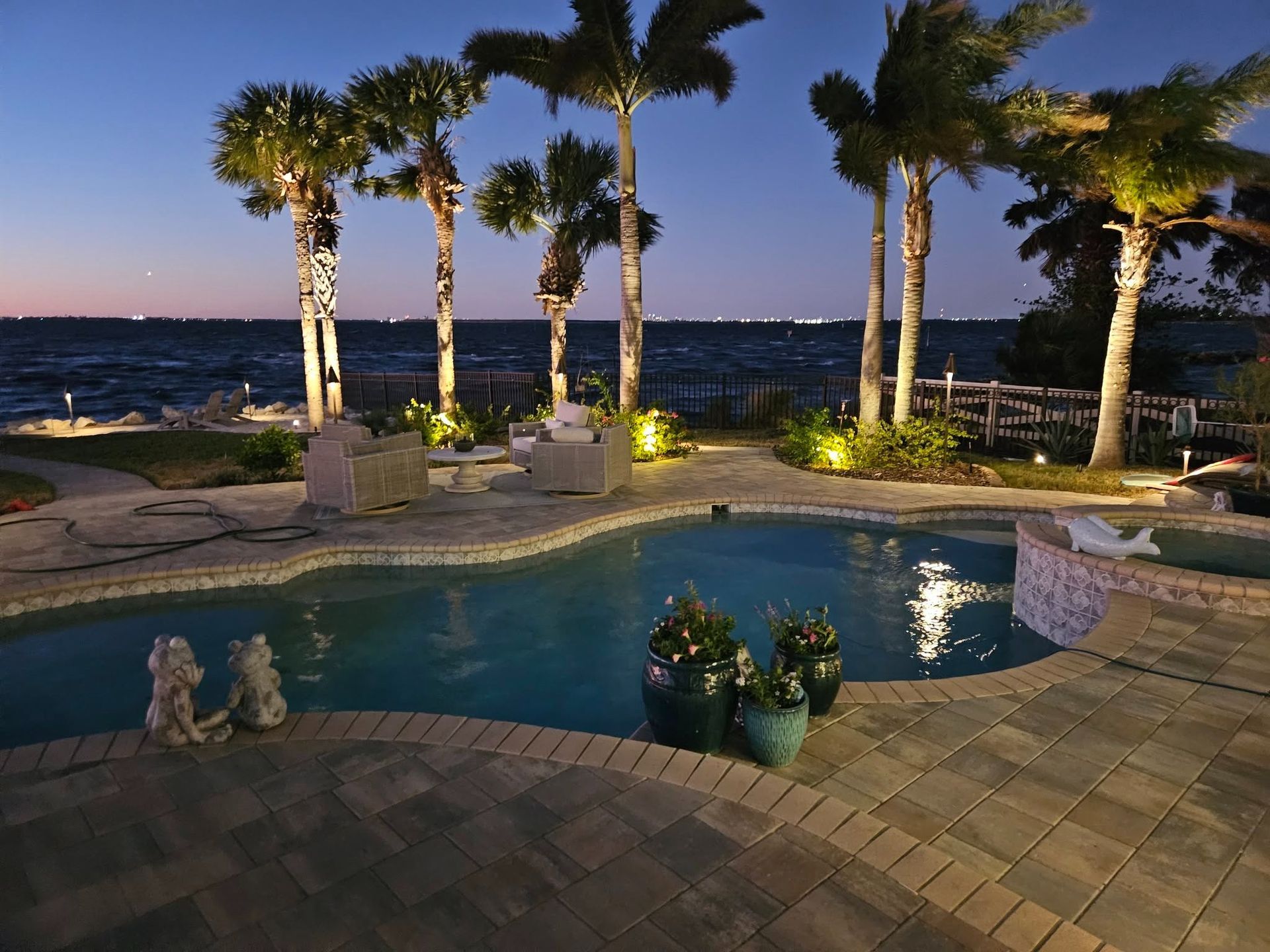 Palm-lined poolside patio at dusk overlooking the ocean, with warm lights and calm water.