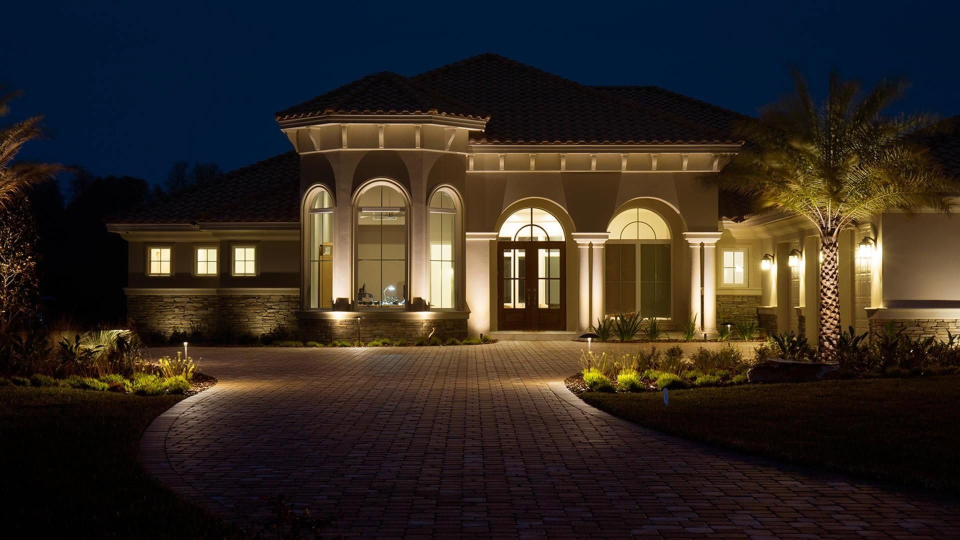 Illuminated stone house at night with arched windows and a lit front entrance behind a dark driveway