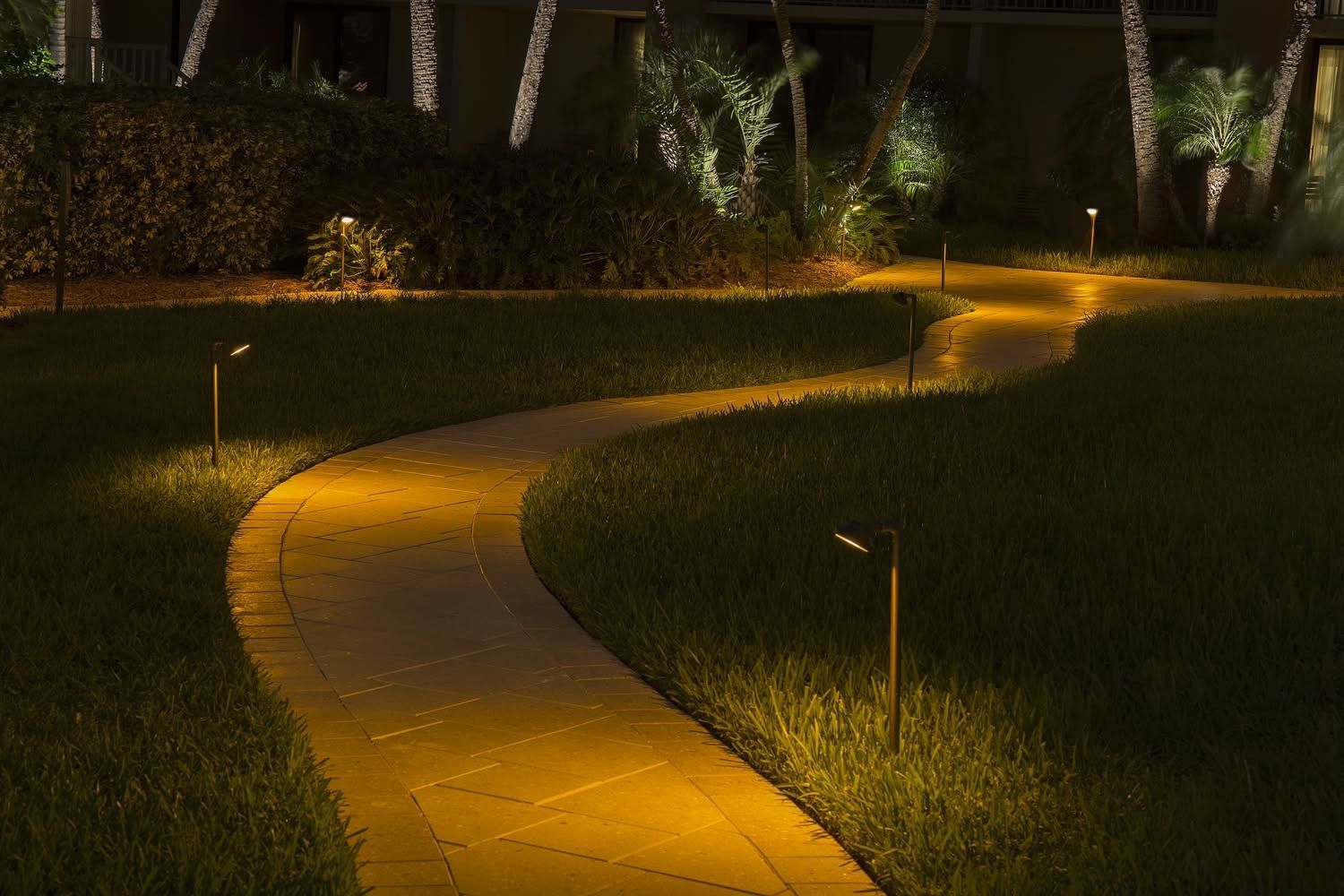 Curved yellow-lit garden pathway at night, bordered by grass and hedges.