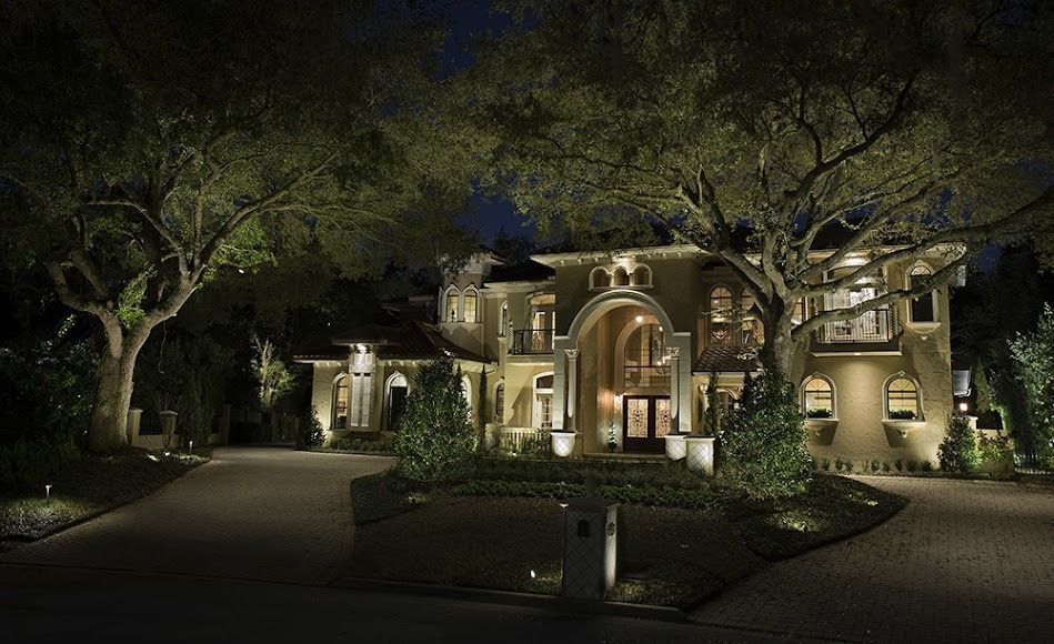 Lit luxury house at night with warm exterior lights, trees, and a curved driveway
