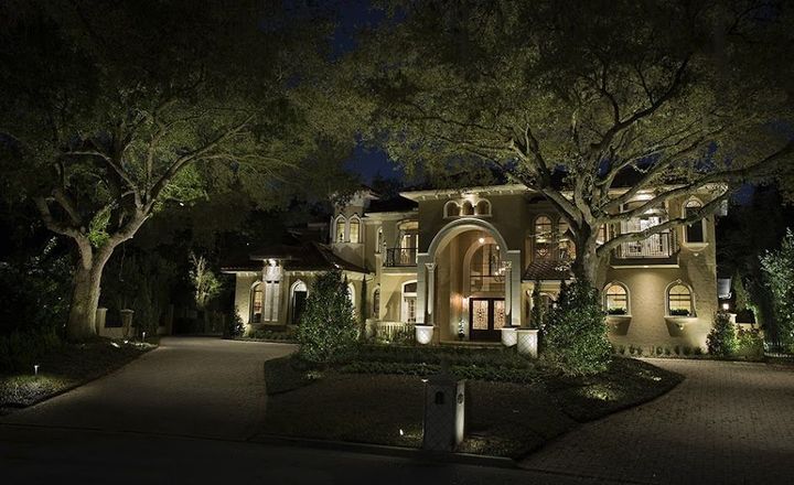 Lit luxury house at night with warm exterior lights, trees, and a curved driveway