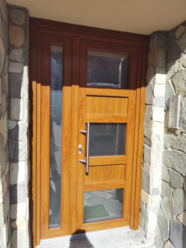 Una porta d'ingresso con finitura effetto legno, pannelli verticali in vetro e maniglia a barra argentata, incastonata in un muro di pietra rustica.