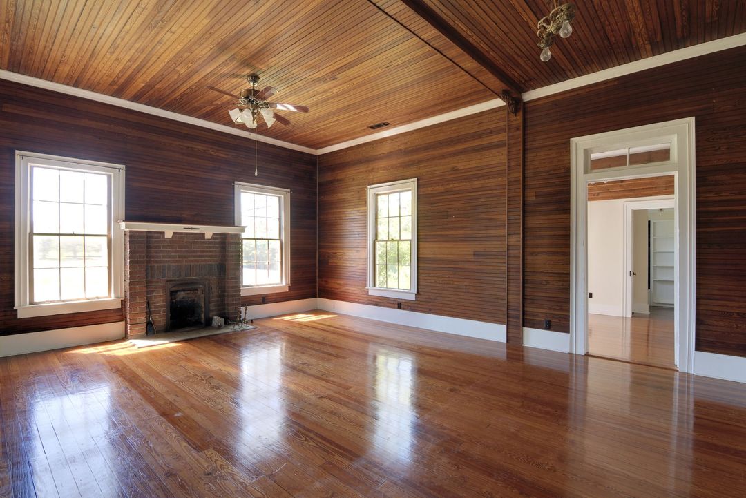 Una stanza spaziosa con pannelli orizzontali in legno scuro, pavimento in parquet, due finestre, camino in mattoni e ventilatori a soffitto.
