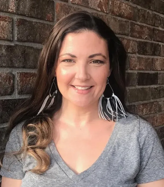 A woman wearing a grey shirt and earrings smiles in front of a brick wall